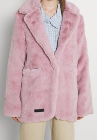 Lichtroze nepbont jas met een sjawlcollar, twee zijzakken en een zwart label aan de onderrand. Gecombineerd met een blauw geruit shirt.