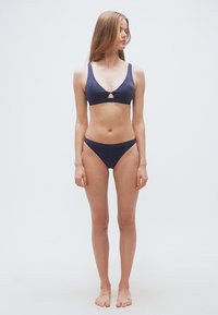 Νέα γυναίκα όρθια ξυπόλητη, φορώντας μπλε μπικίνι με μικρή κοπή στο σουτιέν, κοιτάζοντας προς τα πλάγια σε απλό φόντο.