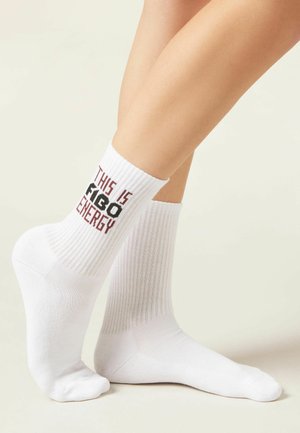 White ribbed crew socks worn on crossed legs, with "THIS IS FIGO ENERGY" text in black and red on the side of one sock.
