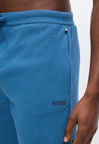 Κοντινό πλάνο ενός ατόμου που φοράει μπλε υφασμάτινο παντελόνι BOSS με ελαστική μέση και ένα χέρι ακουμπισμένο στο πλάι.