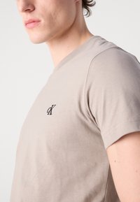 Bežinis trumpomis rankovėmis marškinėliai, pagaminti iš švelnios medvilnės, su mažu juodu logotipu kairėje krūtinės pusėje ir apvaliu kaklu.
