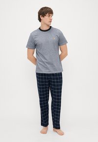 T-shirt en coton gris avec un col noir et un petit logo jaune, associé à un pantalon de pyjama à motifs carreaux navy. Position pieds nus.