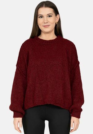 Junge Frau trägt einen locker sitzenden, grob gestrickten, burgunderroten Pullover und schwarze Hosen, steht vor einem schlichten hellen Hintergrund.