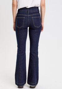 Modelka w ciemnoniebieskich rozszerzanych dżinsach z wysokim stanem, z dwoma tylnymi kieszeniami i kontrastowym żółtym stębnowaniem na szwach.