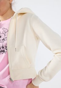 Sudadera de cremallera color crema claro con capucha, bolsillo frontal y puños acanalados. Lleva puesta una camiseta rosa con un diseño gráfico en negro.
