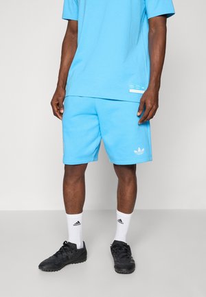 Mann trägt hellblaue Adidas-Shorts und passendes T-Shirt, weiße Adidas-Socken und schwarze Sportschuhe, steht vor einem einfarbigen Hintergrund.