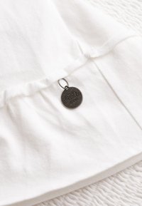 Biała bawełniana bluzka z falbaniastym dołem i srebrną metką z wytłoczonym napisem "Superdry Brand". Miękka tekstura, luźny krój.