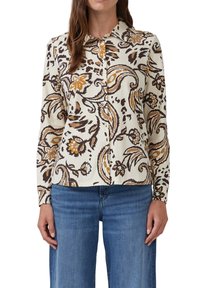 Vrouw in een langwerpige blouse met knopen, met een bruin, beige en oranje bloemen-paisley patroon, gecombineerd met blauwe denim jeans.