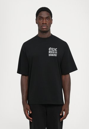 Junger Mann, der ein lockeres schwarzes T-Shirt mit weißem Gothic-Schriftzug "032c Berlin 2001 Hardcore" auf der Brust trägt, vor weißem Hintergrund steht.
