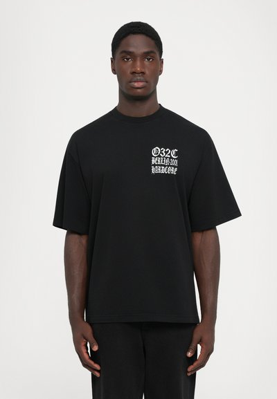 Jonge man die een losse zwarte T-shirt draagt met witte Gothic-stijl tekst "032c Berlin 2001 Hardcore" op de borst, staand tegen een witte achtergrond.