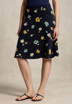 Kvinde iført en marineblå nederdel til knæet med gule, grønne og cremefarvede blomstertryk og sorte flip-flop sandaler, stående på en beige gulv.