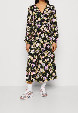 Kobieta ubrana w czarną sukienkę z długim rękawem w różowy, fioletowy i żółty wzór kwiatowy, do tego białe skarpetki i białe trampki.