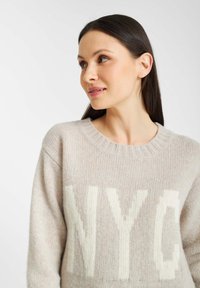 Licht beige gebreide trui met een ronde hals en witte "NYC" opdruk. Zachte textuur en een relaxte pasvorm.