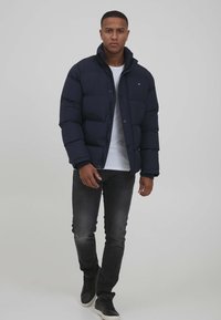 Marineblå puffjakke med høy krage, med glidelås, tykk polstring og glatt tekstur. Brukt over en hvit T-skjorte og mørke jeans.