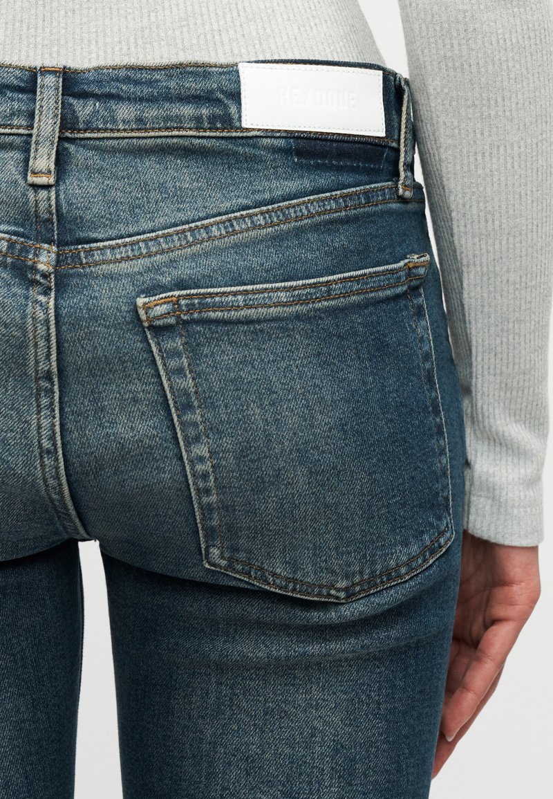 Nahaufnahme einer Person, die blaue Jeans mit einem weißen Markenlabel am Bund und ein graues, geripptes Langarmshirt trägt.
