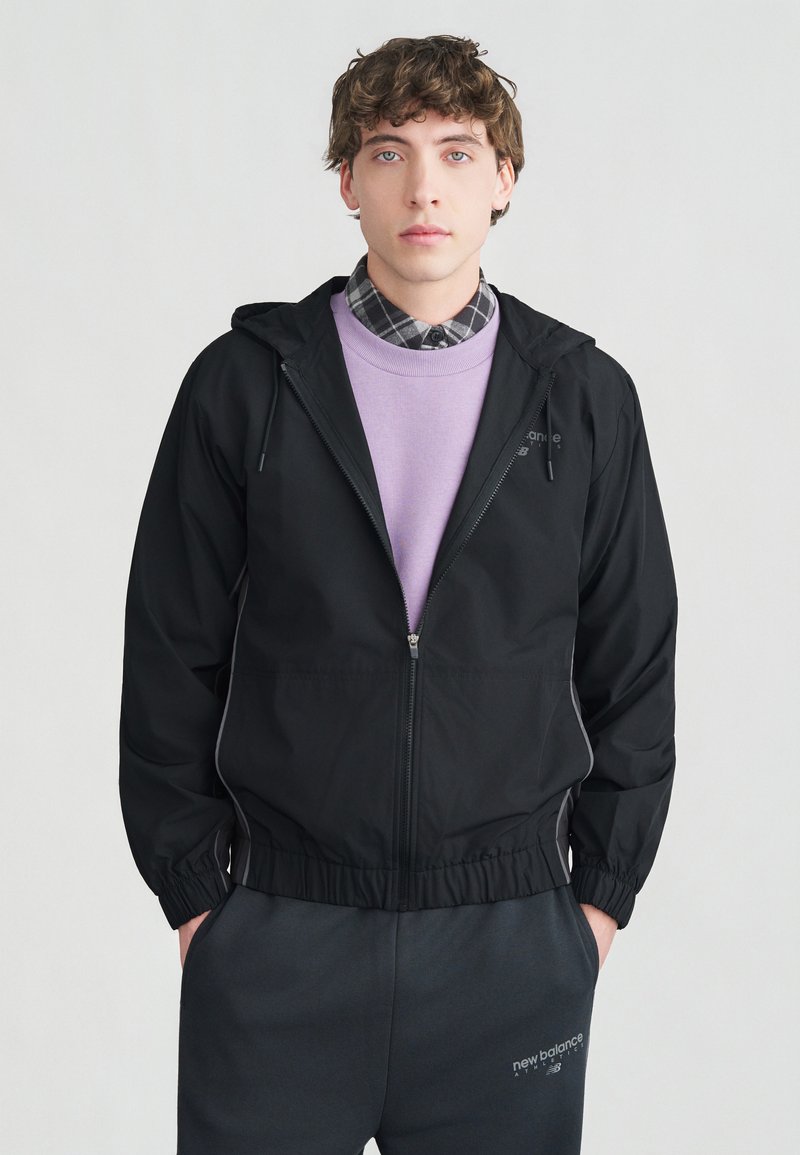 Νεαρός με σγουρά μαλλιά που φοράει μαύρο φούτερ με φερμουάρ και κουκούλα, λιλά πουλόβερ, καρό πουκάμισο και σκούρο παντελόνι New Balance, στέκεται μπροστά σε λευκό φόντο.