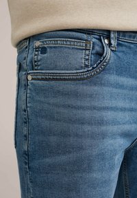 Gaiši zili džinsi ar piecu kabatu dizainu, kas iekļauj vara kniedes un kontrastējošu dzeltenu šūšanu. Gluda tekstūra ar izbalējušu apdari.