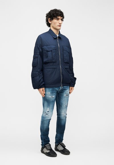 Νεαρός με σγουρά μαλλιά, που φοράει σκούρο μπλε μπουφάν με φερμουάρ και τσέπες, ξεθωριασμένο μπλε τζιν και μαύρα αθλητικά παπούτσια, πάνω σε απλό φωτεινό φόντο.