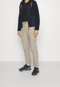Giacca in pile blu navy con cappuccio, pantaloni cargo beige con tasche laterali e scarpe sportive nere con dettagli teal su sfondo bianco.