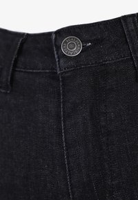 Černé denimové džíny s texturovaným povrchem, viditelným knoflíkem s vyrytým vzorem a čistými švy podél pasu a kapes.