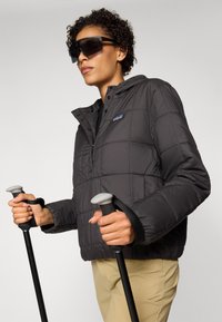 Zwarte gewatteerde pufferjack met capuchon, voorzien van drukknoopsluiting en een logo. Het model houdt trekkingstokken vast met zwarte grips en polsbanden.