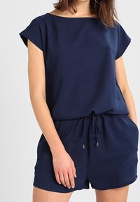 Rompers bleu marine en tissu doux, avec un décolleté bateau, des manches courtes, une taille élastique et un short ample avec des détails subtils.