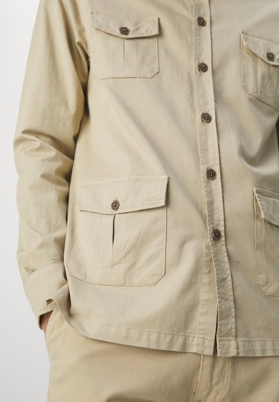 Beige button-up shirt made of fabric with a smooth texture. Features two chest pockets with button closures and a straight hem.