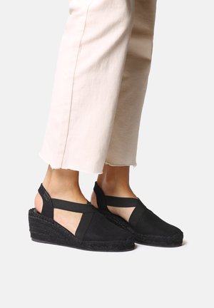 Sandalias de cuña espadrille negras con correas entrecruzadas, parte superior de tela y suela de yute trenzado. Combinadas con pantalones cortos beige claro.