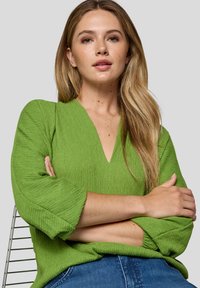 Eine Frau mit langen blonden Haaren, die eine grüne strukturierte V-Ausschnitt-Bluse und Blue Jeans trägt, sitzt mit verschränkten Armen vor einem schlichten Hintergrund.