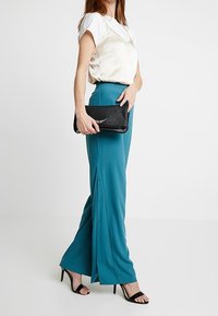 Pantalones de pierna ancha en color verde azulado con una textura suave, con botones laterales. Combinados con una blusa de satén crema y un bolso de mano negro. Tacones negros de tiras.