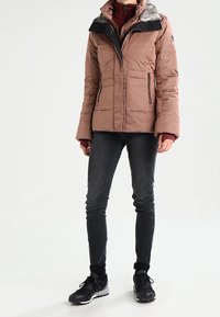 Chaqueta marrón acolchada con cuello de piel sintética negra, bolsillos con cremallera, usada sobre una camiseta granate. Combinada con vaqueros oscuros y zapatillas negras.