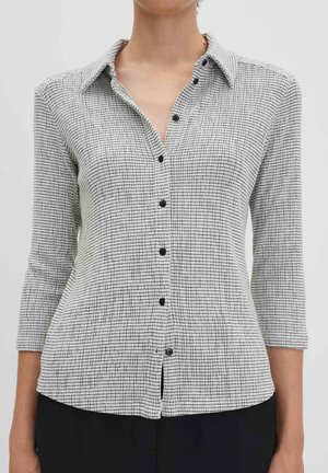 Donna che indossa una camicia grigia a quadri con bottoni e maniche tre quarti, pantaloni neri larghi e corti, e sandali neri con tacco.