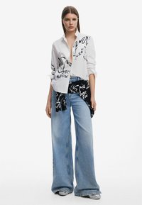 Camicia bianca con motivi astratti neri, abbinata a jeans a gamba larga blu. Un foulard nero legato in vita aggiunge contrasto e dettagli.