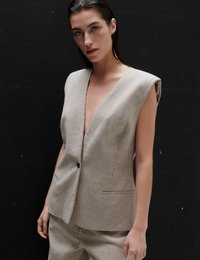 Νέα γυναίκα με βρεγμένα μαλλιά που φοράει αμάνικο σακάκι με βαθύ V λαιμόκοψη, μπεζ ριγέ από ασορτί παντελόνι, σε σκούρο φόντο.