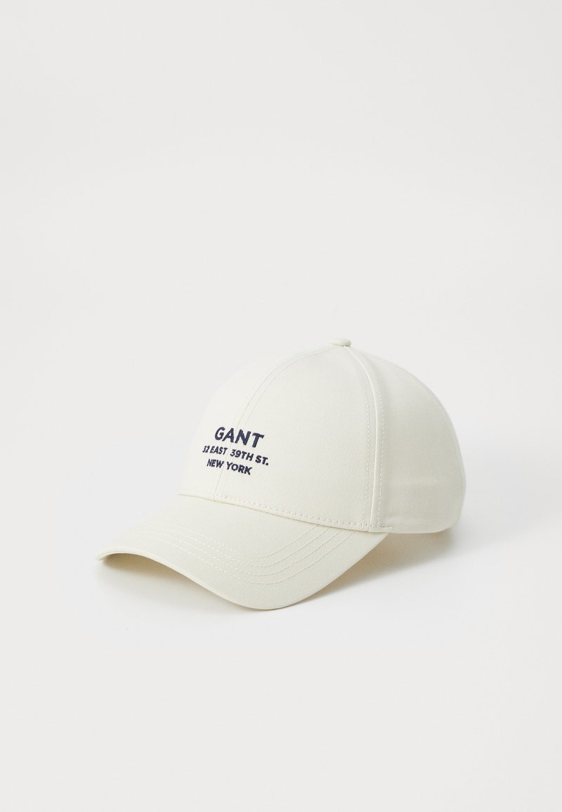 Vit basebollkeps i textil med struktur. Har mörkblå broderad text: "GANT 11 EAST 39TH ST. NEW YORK." Böjd skärm.