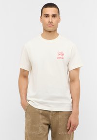 T-shirt en coton écru avec un col rond et des manches courtes, arborant un design graphique rouge sur la poitrine gauche. Assorti à un pantalon beige.