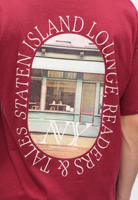 Μαρούλι πουκάμισο με φωτογραφία καταστήματος περιφραγμένη από λευκό κείμενο που λέει "Staten Island Lounge Readers & Tales" και λογότυπο "Originals Studio".