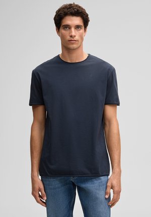 Jonge man staat recht voor zich uit kijkend, draagt een effen donkerblauw T-shirt met korte mouwen en een blauwe spijkerbroek, tegen een grijze achtergrond.