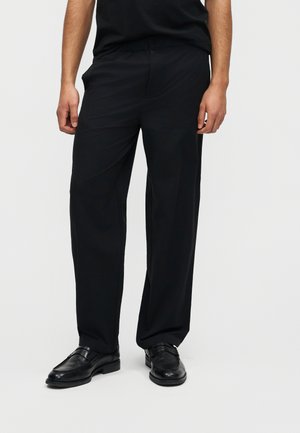 Άνδρας που φοράει μαύρο παντελόνι με φαρδιά πόδια και μαύρα δερμάτινα επίσημα παπούτσια, στέκεται μπροστά σε απλό ανοιχτόχρωμο φόντο.