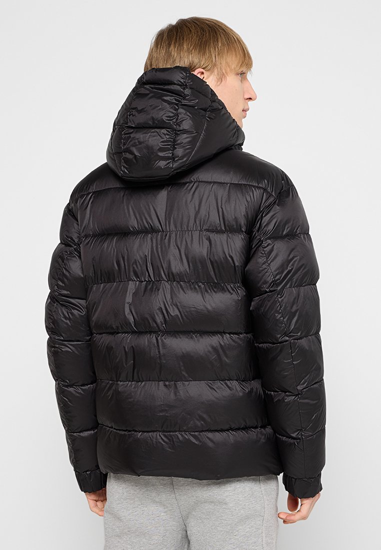 Blonde Person, die eine schwarze Daunenjacke mit Kapuze trägt, seitlich blickend, kombiniert mit hellgrauen Hosen vor einem schlichten Hintergrund.