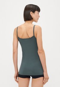 Donkergroene tanktop gemaakt van zacht materiaal, heeft dunne bandjes en een gladde textuur. Ggedragen met marineblauwe shorts. Achteraanzicht weergegeven.
