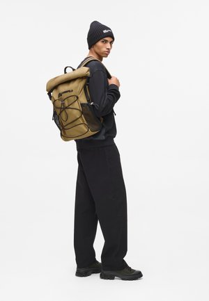 Khaki rull-funktionsryggsäck med svart nätduk, justerbara axelremmar och dragsnöre stängning. Modell bär en svart outfit och mössa.