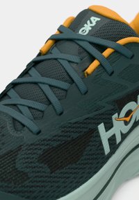 Chaussure de course avec une tige en mesh vert foncé à motif camouflage, accents orange, et semelle épaisse. Comprend des lacets et un logo bleu sarcelle.