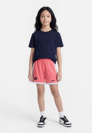 Joven de pie mirando hacia adelante, vistiendo una camiseta azul marino, pantalones cortos deportivos coral con logotipos, calcetines blancos y zapatillas negras y blancas.