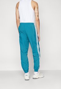 Uomo che indossa pantaloni sportivi teal con strisce bianche, canotta bianca, calze bianche e sneaker Adidas bianche, in piedi davanti a uno sfondo semplice.