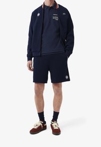 Marinblå idrottsklädsel som inkluderar en zip-up jacka, en polo med klubbens logo, matchande shorts och bruna sneakers med vita snörningar.