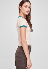 Beige short-sleeve t-shirt with teal trim, fitted design, smooth texture. Paired with dark brown high-waisted pants with pockets.