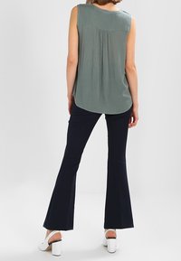 Haut sans manches vert clair avec des plis dans le dos ; associé à un pantalon évasé bleu marine. Chaussures : sandales blanches à talons.