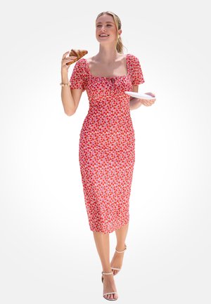 Kobieta w czerwonej sukience w wzór wiśni trzyma talerz i je rogalika, uśmiechając się, stojąc na białym tle.