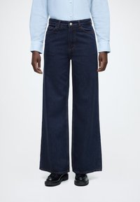 Modré rifle širokého strihu z denimu s vysokým pásom, piatimi vreckami a kontrastným prešívaním; skombinované s čiernymi topánkami.